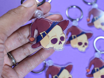 Keychain with skull design wearing a red hat held by a hand against a purple background with more keychains.