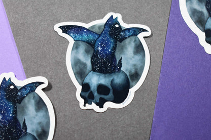 Sticker of a skull with a cat with bat wings sitting on it with a galaxy design on a gray and purple background
