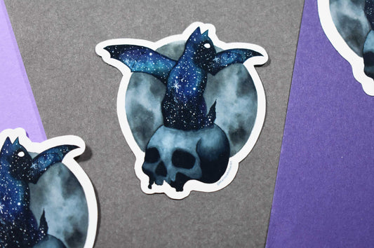 Sticker of a skull with a cat with bat wings sitting on it with a galaxy design on a gray and purple background