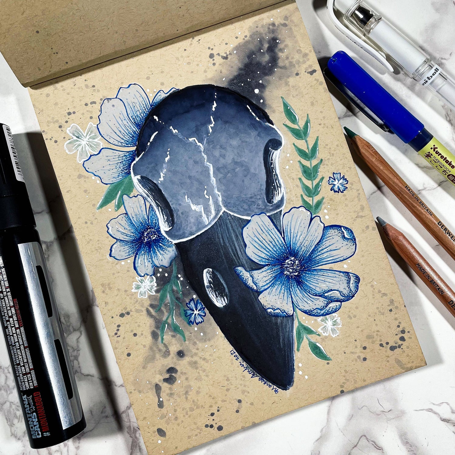 Artistic depiction of a raven skull with blue flowers on a textured surface, surrounded by art supplies.