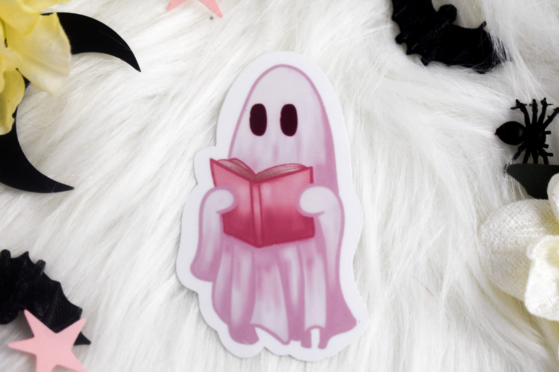 Ghost character holding a pink book on a textured white background with Halloween decorations.