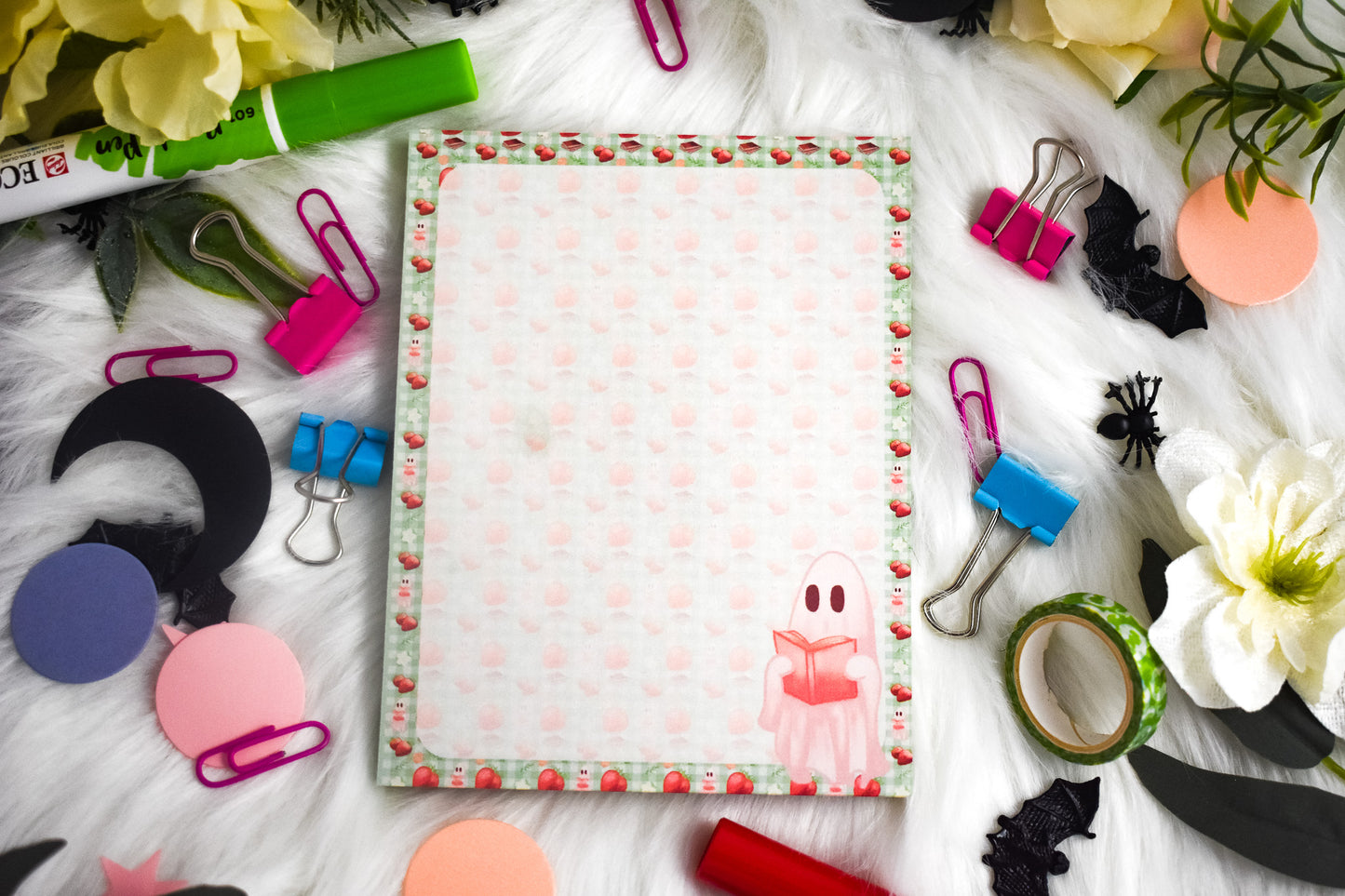 Notepad with colorful strawberry and ghost design surrounded by stationery items on a white surface