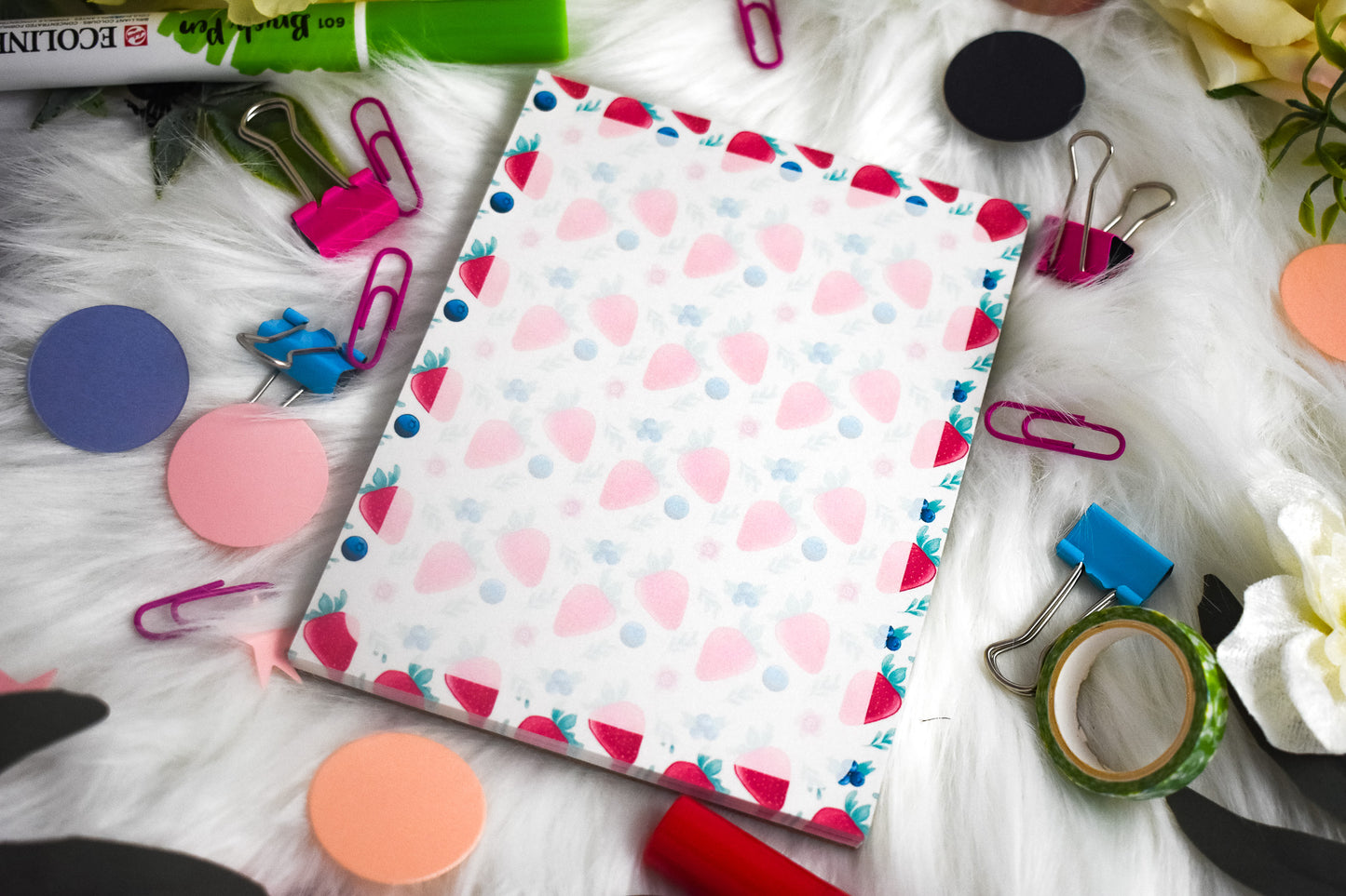 Notepad with colorful strawberry design surrounded by stationery items on a white surface