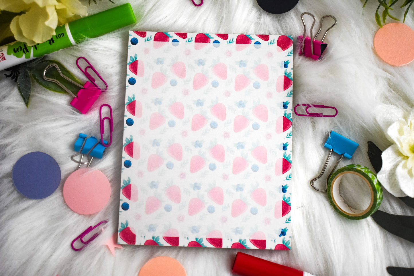 Notepad with colorful strawberry design surrounded by stationery items on a white surface