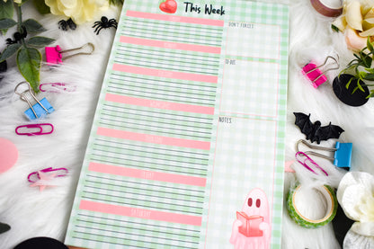Weekly planner pad with Halloween-themed designs on a decorative surface with stationery items.