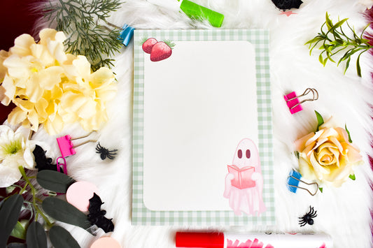 Notepad with a ghost illustration on a desk with flowers and stationery items