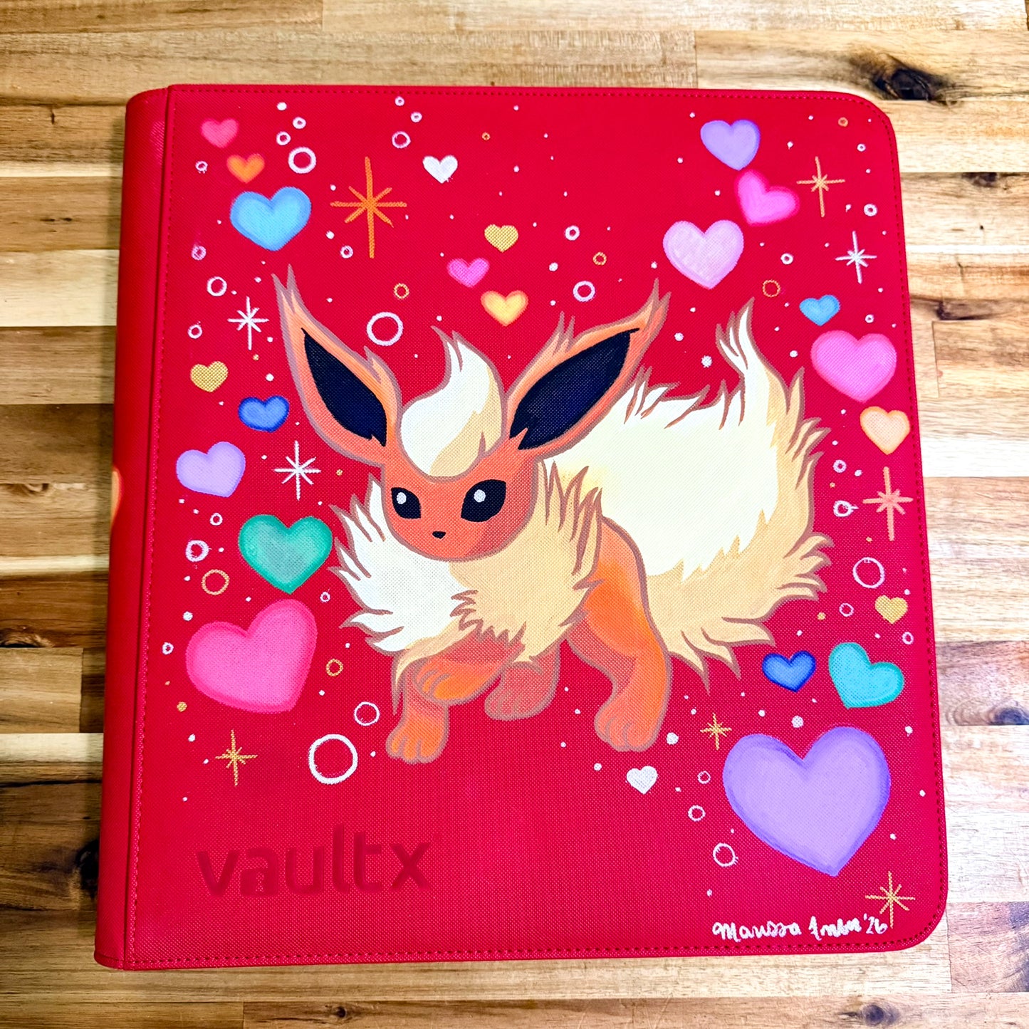 A red vinyl binder cover featuring a painted illustration of a fluffy cream and orange fox-like character, surrounded by colorful hearts and sparkles. The word 'vaultx' is at the bottom left, and a small artist signature is visible in the bottom right corner.
