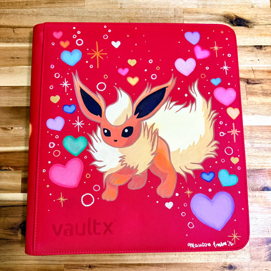 A red vinyl binder cover featuring a painted illustration of a fluffy cream and orange fox-like character, surrounded by colorful hearts and sparkles. The word 'vaultx' is at the bottom left, and a small artist signature is visible in the bottom right corner.