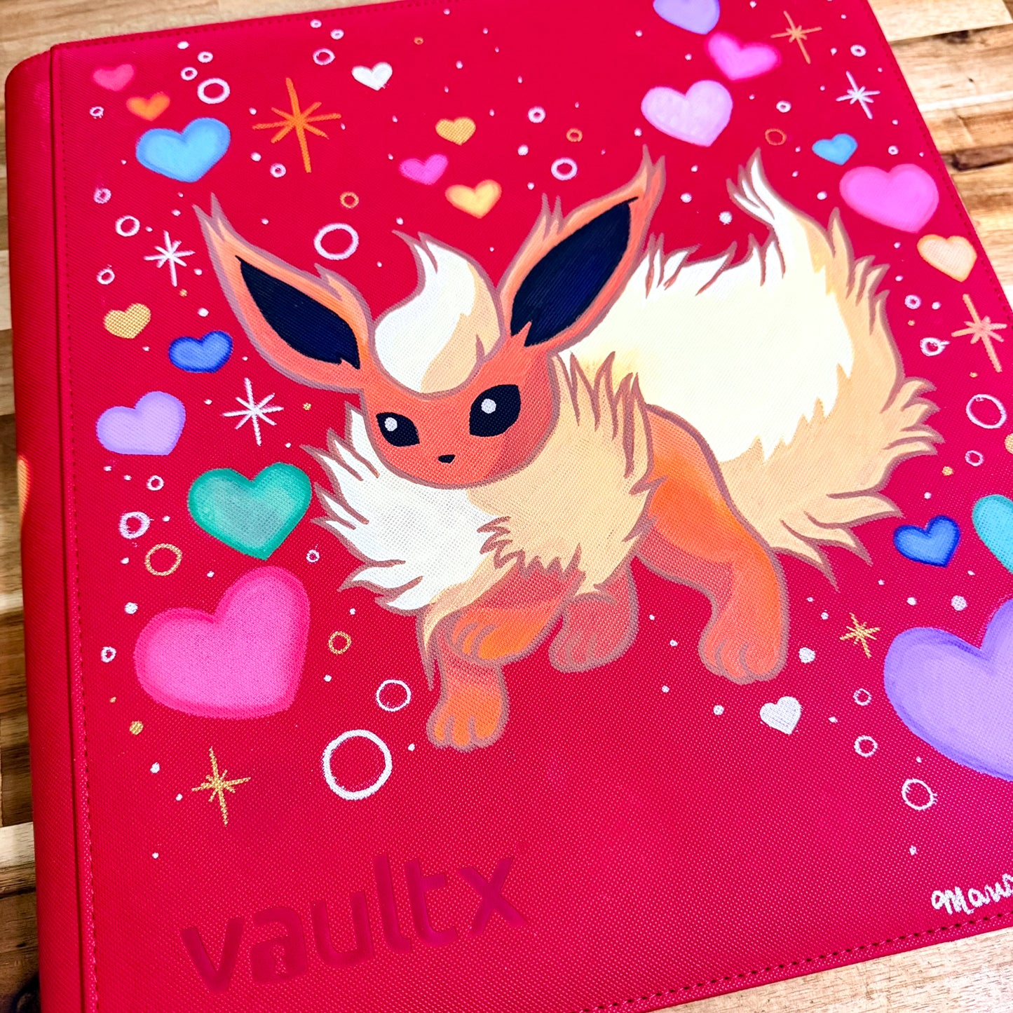 A red vinyl binder cover featuring a painted illustration of a fluffy cream and orange fox-like character, surrounded by colorful hearts and sparkles. The word 'vaultx' is at the bottom left, and a small artist signature is visible in the bottom right corner.