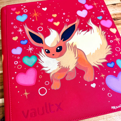 A red vinyl binder cover featuring a painted illustration of a fluffy cream and orange fox-like character, surrounded by colorful hearts and sparkles. The word 'vaultx' is at the bottom left, and a small artist signature is visible in the bottom right corner.