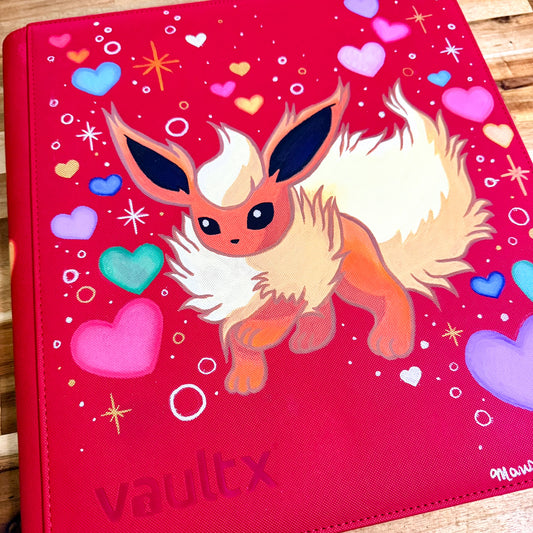 A red vinyl binder cover featuring a painted illustration of a fluffy cream and orange fox-like character, surrounded by colorful hearts and sparkles. The word 'vaultx' is at the bottom left, and a small artist signature is visible in the bottom right corner.