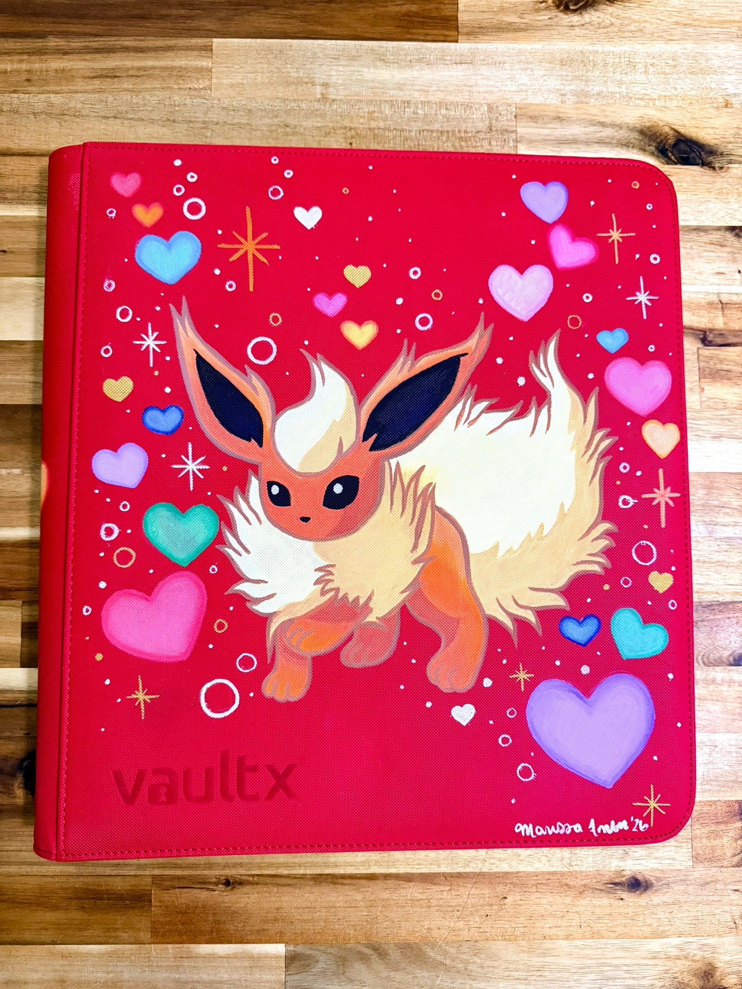 A red vinyl binder cover featuring a painted illustration of a fluffy cream and orange fox-like character, surrounded by colorful hearts and sparkles. The word 'vaultx' is at the bottom left, and a small artist signature is visible in the bottom right corner.