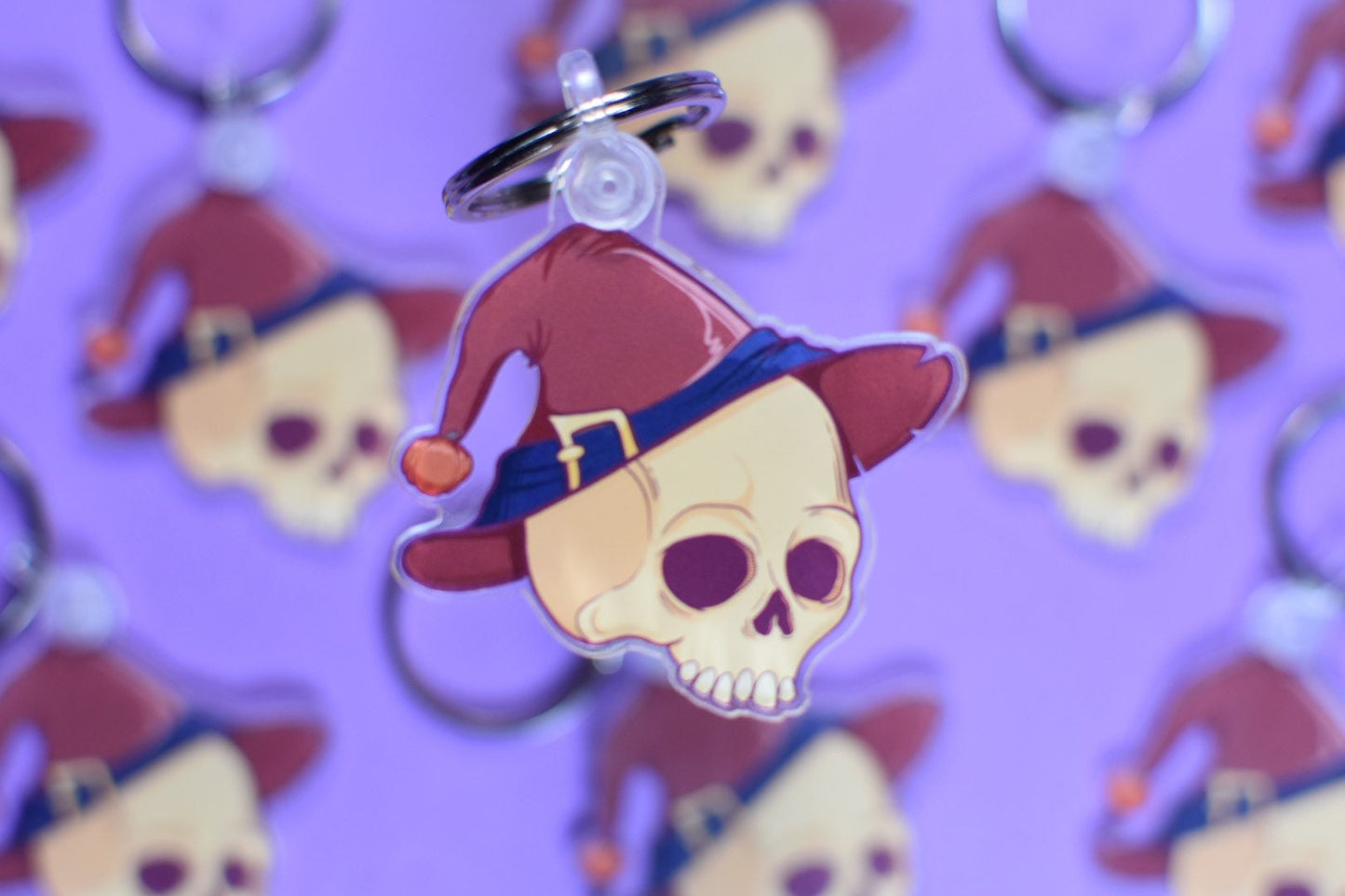Keychain with skull design wearing a red witches hat on a purple background with similar keychains.
